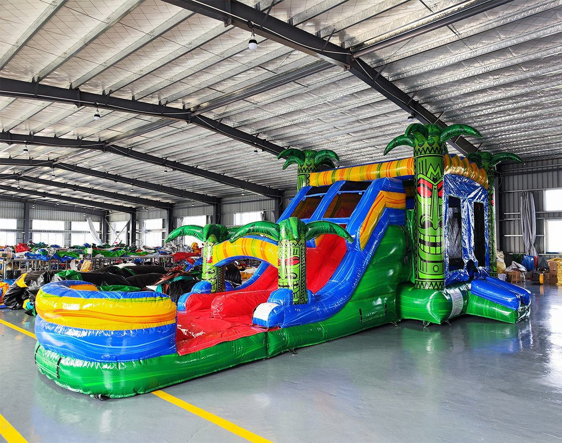 Three-quarter view of Reggae Rush Dual Lane Commercial Inflatable Combo in a warehouse environment. The inflatable features green palm trees, dual blue slides, a yellow and green bounce house area, and a vibrant splash pool. Other inflatable products and warehouse materials in the background demonstrate its commercial application.