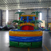 Front view of Reggae Rush Dual Lane Commercial Inflatable Combo (Wet/Dry) set up in a large indoor warehouse. The inflatable features a tropical theme with green palm tree pillars, dual blue slides, a yellow and green bounce house area with mesh windows, and a colorful splash pool base. The structure has a vibrant design with red, yellow, green, and blue colors. Various other inflatable products and warehouse equipment are visible in the background, showcasing the industrial setting with metal roofing.