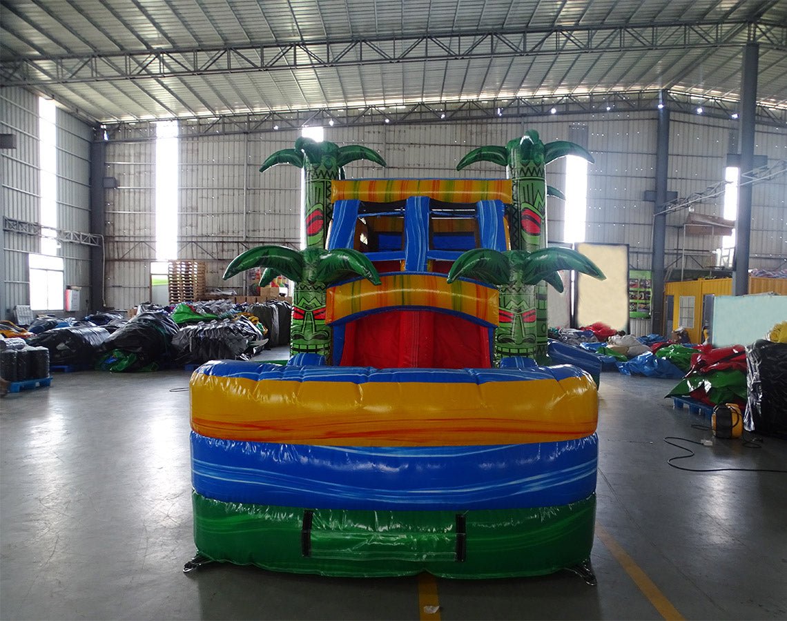 Front view of Reggae Rush Dual Lane Commercial Inflatable Combo (Wet/Dry) set up in a large indoor warehouse. The inflatable features a tropical theme with green palm tree pillars, dual blue slides, a yellow and green bounce house area with mesh windows, and a colorful splash pool base. The structure has a vibrant design with red, yellow, green, and blue colors. Various other inflatable products and warehouse equipment are visible in the background, showcasing the industrial setting with metal roofing.