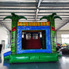 Rear view of Reggae Rush Dual Lane Commercial Inflatable Combo set up in a large indoor warehouse. The inflatable features green palm tree pillars, dual blue slides, a yellow and green bounce house area with mesh windows, and a colorful splash pool. Metal roofing and various other inflatable products are visible in the background.