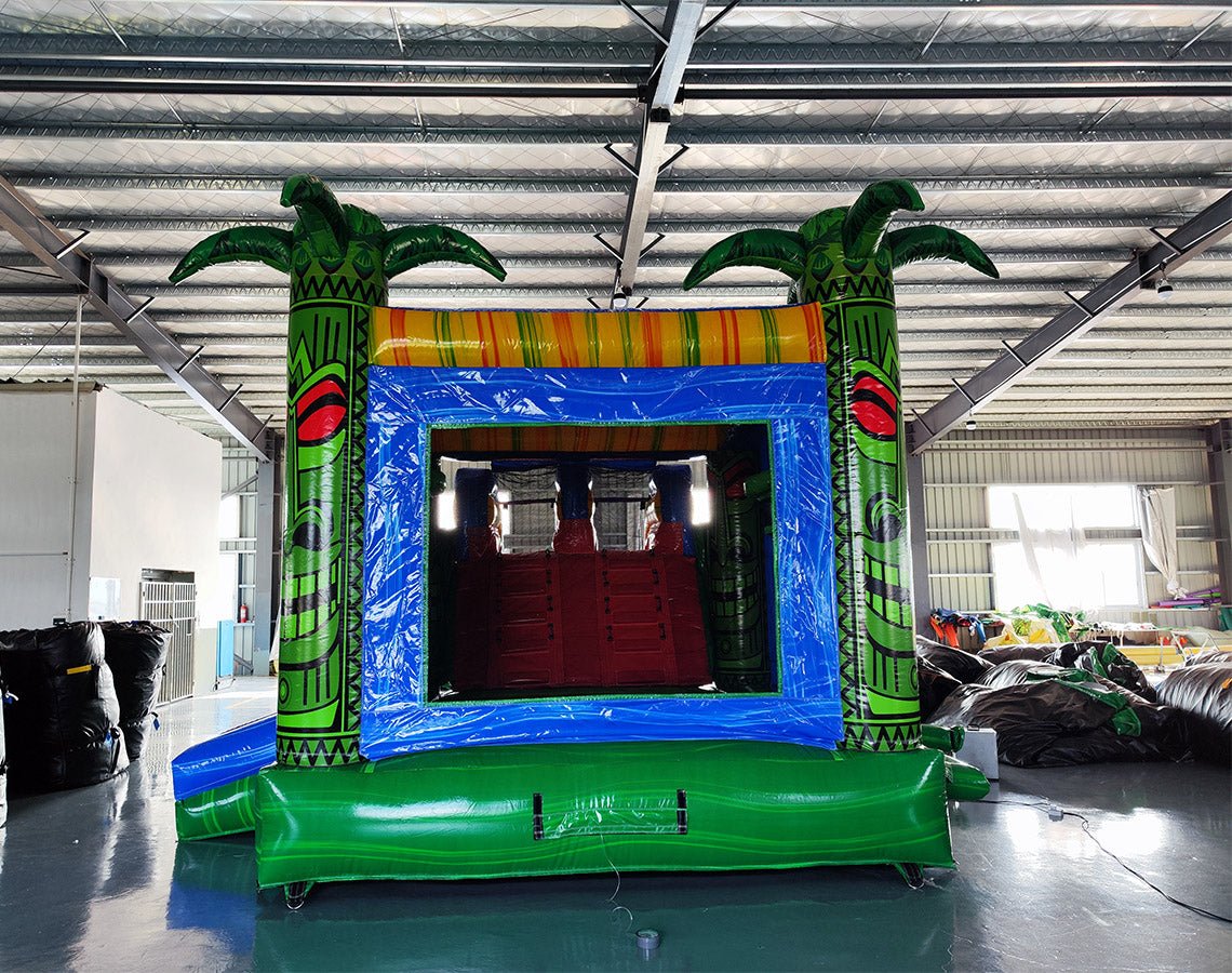 Rear view of Reggae Rush Dual Lane Commercial Inflatable Combo set up in a large indoor warehouse. The inflatable features green palm tree pillars, dual blue slides, a yellow and green bounce house area with mesh windows, and a colorful splash pool. Metal roofing and various other inflatable products are visible in the background.