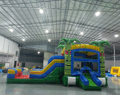 Wide-angle view of Reggae Rush Commercial Econo Inflatable Combo (Wet/Dry) in an indoor warehouse setting. The inflatable features tiki face designs, palm tree decorations, yellow slide, and blue splash pool. Other inflatable products, concrete flooring, and warehouse walls visible, demonstrating its size and commercial setting.