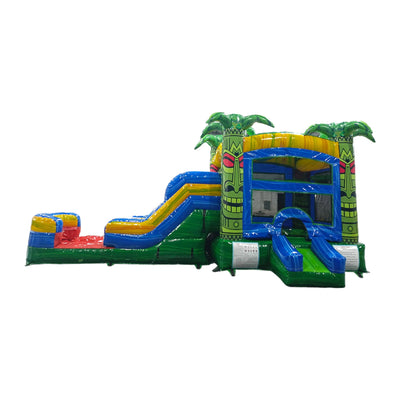 Large inflatable tropical-themed combo featuring a bounce house with tiki face designs, palm tree decorations, attached slide, and splash pool. The structure has vibrant green, yellow, blue, and red colors with rasta-inspired accents. Tiki totems flank the entrance, and inflatable palm trees top the structure. Isolated on a white background.