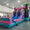 Three-quarter view of Retro Splash Dual Lane Commercial Inflatable Combo in a warehouse setting. The structure displays a pink and blue retro van design, dual blue slides, inflatable palm trees, and a large splash pool area. Various other inflatable products and warehouse equipment are visible, demonstrating its size and commercial application.