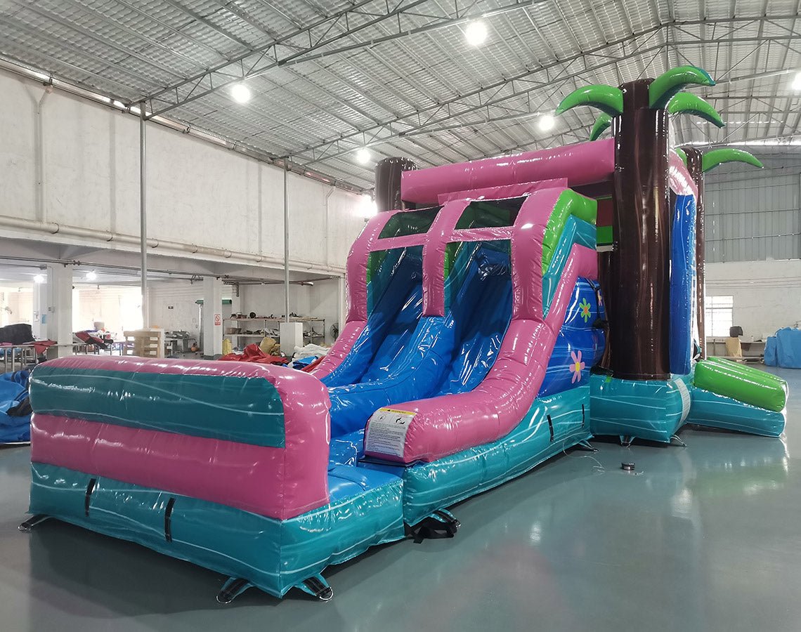 Three-quarter view of Retro Splash Dual Lane Commercial Inflatable Combo in a warehouse setting. The structure displays a pink and blue retro van design, dual blue slides, inflatable palm trees, and a large splash pool area. Various other inflatable products and warehouse equipment are visible, demonstrating its size and commercial application.