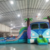 Cross side view of Retro Splash Dual Lane Commercial Inflatable Combo (Wet/Dry) featuring a vintage van design. The structure has dual blue slides, a pink and blue van-shaped bounce house area with a peace sign, inflatable palm trees, and a surfboard. A white seagull figure sits atop the van. Set up in a large indoor warehouse with metal roofing and bright lighting visible.