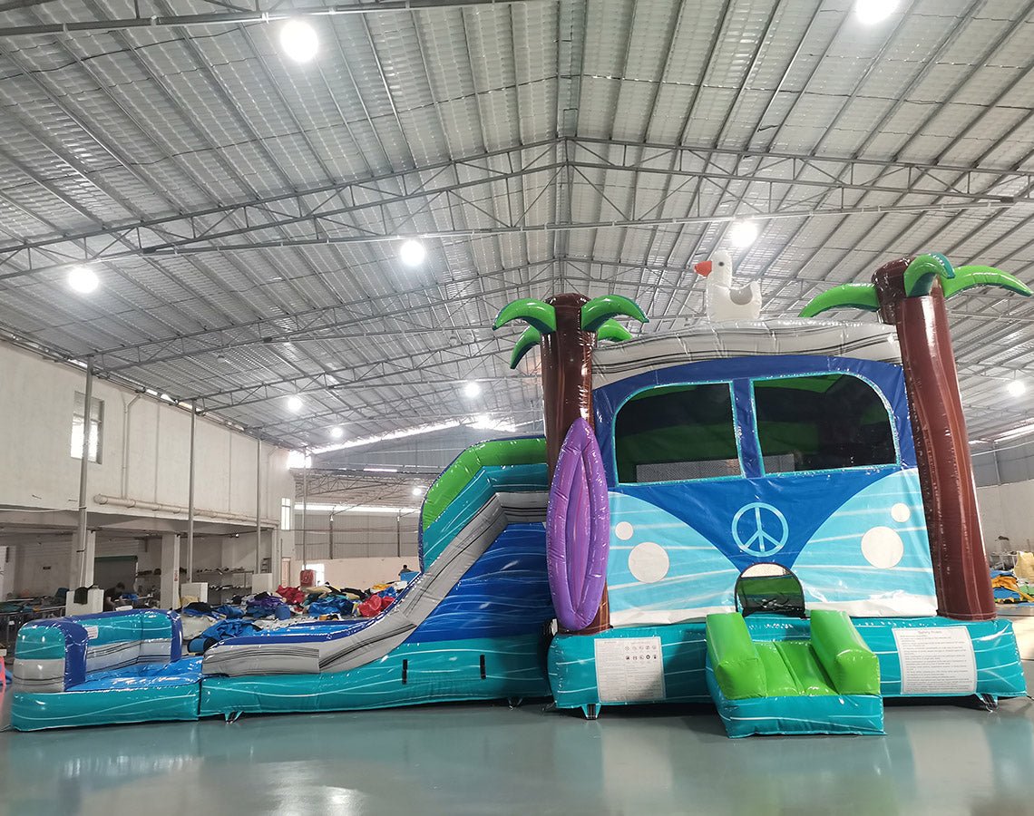 Cross side view of Retro Splash Dual Lane Commercial Inflatable Combo (Wet/Dry) featuring a vintage van design. The structure has dual blue slides, a pink and blue van-shaped bounce house area with a peace sign, inflatable palm trees, and a surfboard. A white seagull figure sits atop the van. Set up in a large indoor warehouse with metal roofing and bright lighting visible.