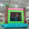 Rear view of Retro Splash Dual Lane Commercial Inflatable Combo (Wet/Dry) showing the entrance area. Features a green bounce house frame with brown inflatable palm tree trunks and green leaves. A pink canopy tops the structure. Set in a warehouse with concrete flooring and various other inflatable products visible in the background.