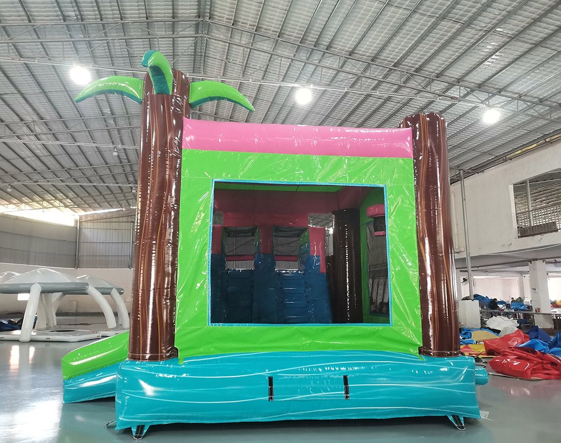 Rear view of Retro Splash Dual Lane Commercial Inflatable Combo (Wet/Dry) showing the entrance area. Features a green bounce house frame with brown inflatable palm tree trunks and green leaves. A pink canopy tops the structure. Set in a warehouse with concrete flooring and various other inflatable products visible in the background.