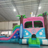 Three-quarter view of Retro Splash Dual Lane Commercial Inflatable Combo (Wet/Dry) set up in a warehouse. The inflatable features a pink and blue retro van design with a peace sign, dual blue slides, inflatable palm trees, a surfboard, and a white seagull on top. A blue splash pool area and green entrance ramp are visible. Various other inflatable products and warehouse equipment can be seen in the background.