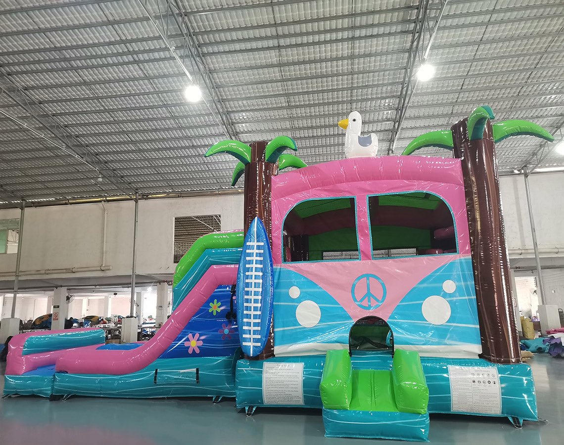 Three-quarter view of Retro Splash Dual Lane Commercial Inflatable Combo (Wet/Dry) set up in a warehouse. The inflatable features a pink and blue retro van design with a peace sign, dual blue slides, inflatable palm trees, a surfboard, and a white seagull on top. A blue splash pool area and green entrance ramp are visible. Various other inflatable products and warehouse equipment can be seen in the background.