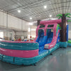 Wide-angle view of Retro Splash Dual Lane Commercial Inflatable Combo set up in a large indoor warehouse. The inflatable features a pink and blue retro van design, dual blue slides, inflatable palm trees, and a large splash pool area. Metal roofing, bright lighting, and various other inflatable products are visible in the background.