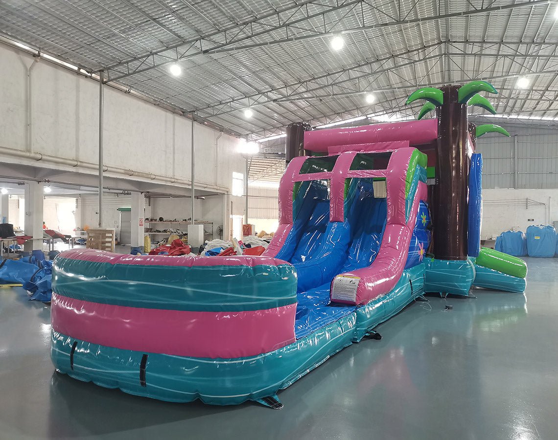 Wide-angle view of Retro Splash Dual Lane Commercial Inflatable Combo set up in a large indoor warehouse. The inflatable features a pink and blue retro van design, dual blue slides, inflatable palm trees, and a large splash pool area. Metal roofing, bright lighting, and various other inflatable products are visible in the background.