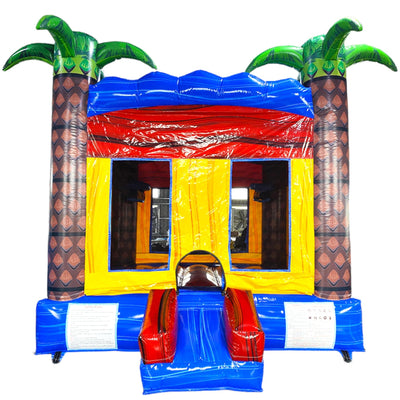 Tropical-themed inflatable Rip Curl Commercial Grade Bounce House featuring a vibrant design with two large inflatable palm trees flanking a blue and yellow bounce area. The structure has a red roof with wave-like patterns, yellow walls with mesh windows, and a small blue slide at the entrance. Set up in a spacious indoor warehouse with metal roofing and fluorescent lighting visible.