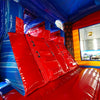 Interior view of Rip Curl Dual Lane Commercial Inflatable Combo (Wet/Dry) showing dual red slides with blue safety barriers. The inflatable features a blue ceiling, yellow side walls, and a colorful tropical-themed backdrop visible through the exit. Set up in a large indoor warehouse with metal roofing and bright lighting visible above.
