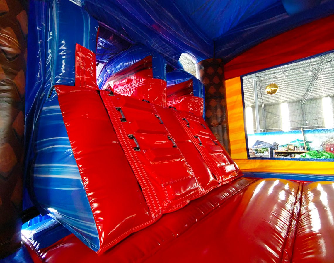 Interior view of Rip Curl Dual Lane Commercial Inflatable Combo (Wet/Dry) showing dual red slides with blue safety barriers. The inflatable features a blue ceiling, yellow side walls, and a colorful tropical-themed backdrop visible through the exit. Set up in a large indoor warehouse with metal roofing and bright lighting visible above.
