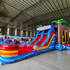 Wide-angle view of Rip Curl Dual Lane Commercial Inflatable Combo (Wet/Dry) set up in a large indoor warehouse. The inflatable displays dual blue slides, brown palm tree trunks with blue leaves, a yellow and blue bounce house area, and a large circular splash pool with rainbow-colored rings. Various other inflatable products, storage containers, and warehouse equipment are visible, showcasing its size and commercial setting.
