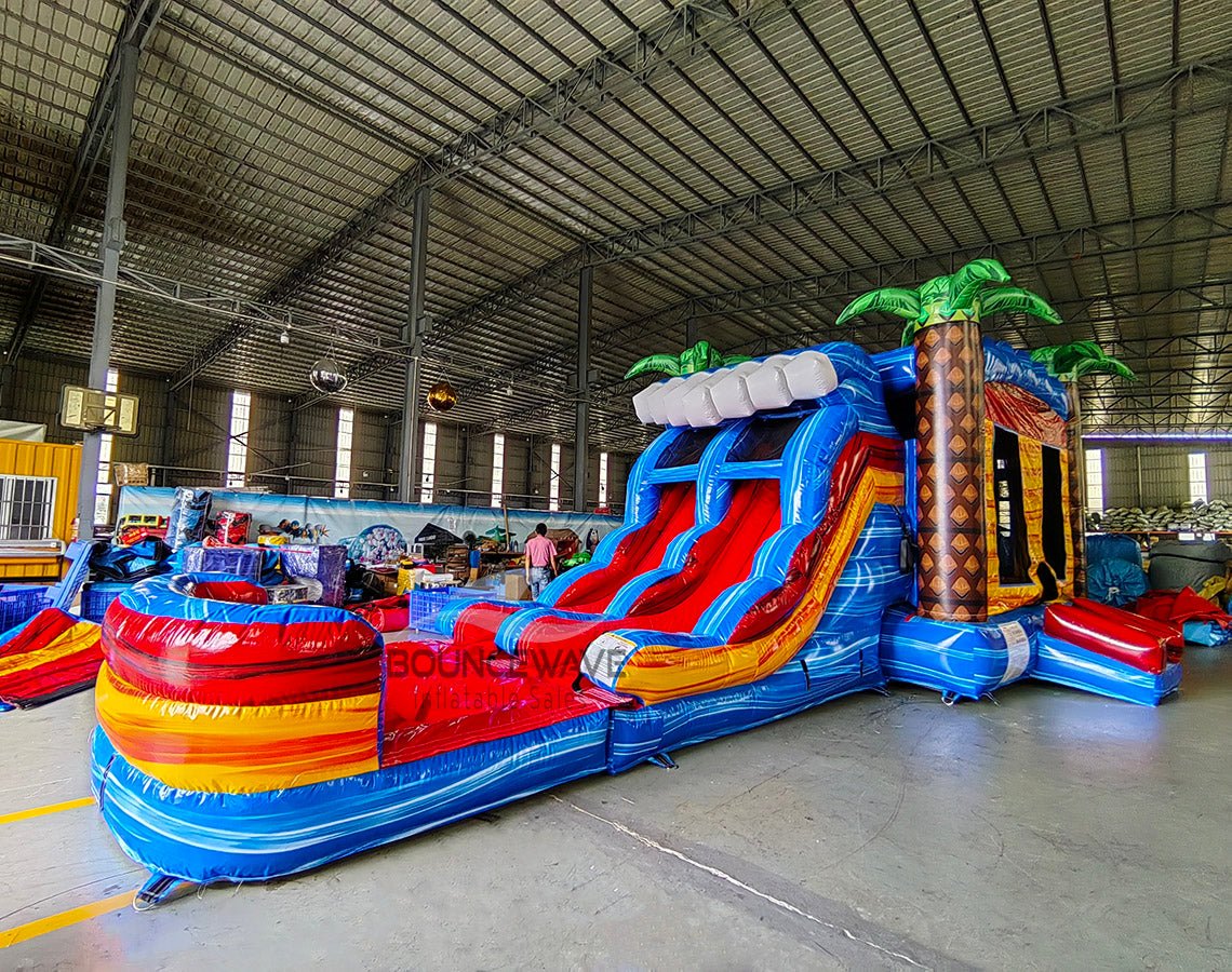 Wide-angle view of Rip Curl Dual Lane Commercial Inflatable Combo (Wet/Dry) set up in a large indoor warehouse. The inflatable displays dual blue slides, brown palm tree trunks with blue leaves, a yellow and blue bounce house area, and a large circular splash pool with rainbow-colored rings. Various other inflatable products, storage containers, and warehouse equipment are visible, showcasing its size and commercial setting.
