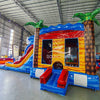 Side view of Rip Curl Dual Lane Commercial Inflatable Combo (Wet/Dry) showing its full length. The inflatable features brown palm tree trunks with blue leaves, dual blue slides, a yellow and blue bounce house structure, and a large circular splash pool with rainbow-colored rings. Set in a large indoor warehouse with metal roofing, concrete flooring, and various other inflatable products visible in the background.
