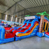 Wide-angle view of Rip Curl Dual Lane Commercial Inflatable Combo (Wet/Dry) in a spacious warehouse. The inflatable displays brown palm tree trunks with green leaves, dual blue slides, a bounce house area, and a large circular splash pool with vibrant rainbow-colored rings. Various other inflatable products, storage containers, and warehouse equipment are visible, showcasing its size and commercial setting.
