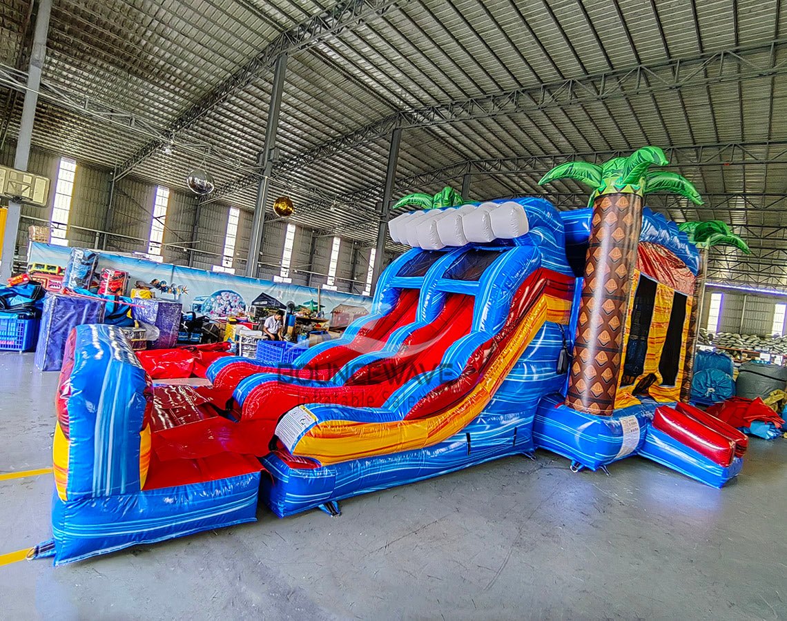 Wide-angle view of Rip Curl Dual Lane Commercial Inflatable Combo (Wet/Dry) in a spacious warehouse. The inflatable displays brown palm tree trunks with green leaves, dual blue slides, a bounce house area, and a large circular splash pool with vibrant rainbow-colored rings. Various other inflatable products, storage containers, and warehouse equipment are visible, showcasing its size and commercial setting.
