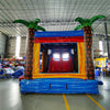 Rear view of Rip Curl Dual Lane Commercial Inflatable Combo (Wet/Dry) set up in a large indoor warehouse. The inflatable features brown palm tree trunks with green leaves, dual entrances, and a colorful base with blue, yellow, orange, and red stripes. 
