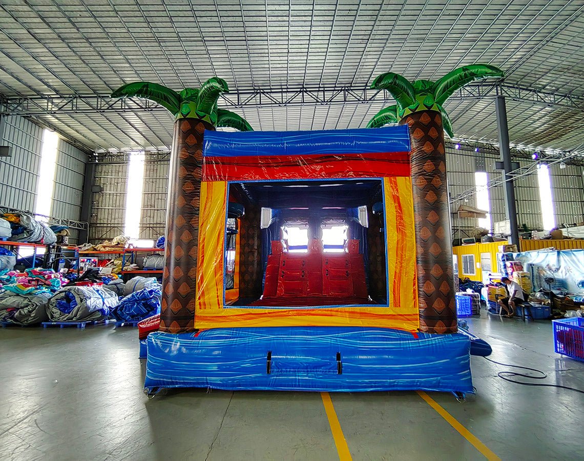 Rear view of Rip Curl Dual Lane Commercial Inflatable Combo (Wet/Dry) set up in a large indoor warehouse. The inflatable features brown palm tree trunks with green leaves, dual entrances, and a colorful base with blue, yellow, orange, and red stripes. 
