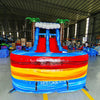 Front view of Rip Curl Dual Lane Commercial Inflatable Combo (Wet/Dry) featuring a tropical theme with inflatable palm trees. The structure has dual blue slides, a yellow and blue bounce house area, and a large circular splash pool with vibrant rainbow-colored rings. Set up in a spacious warehouse with metal roofing and bright lighting visible.
