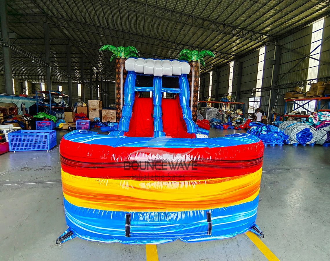 Front view of Rip Curl Dual Lane Commercial Inflatable Combo (Wet/Dry) featuring a tropical theme with inflatable palm trees. The structure has dual blue slides, a yellow and blue bounce house area, and a large circular splash pool with vibrant rainbow-colored rings. Set up in a spacious warehouse with metal roofing and bright lighting visible.
