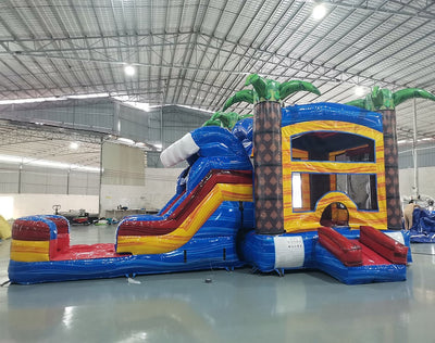 Wide-angle view of Rip Curl Econo Combo featuring a tropical-themed bounce house with palm tree decorations, curved slide, and large splash pool. The inflatable has bright blue, yellow, and red colors and is set up in a spacious indoor warehouse with metal roofing and other equipment visible.