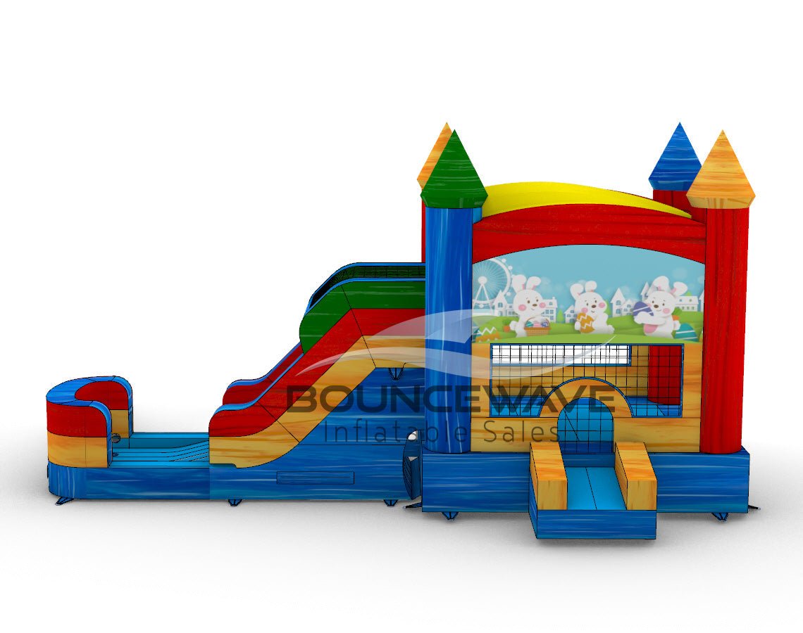 Side view of Seasonal Sampler Commercial Econo Inflatable Panel Combo featuring a colorful castle-like structure with multiple towers and spires. The inflatable has dual slides, one red and one blue, emerging from a yellow and red base. A bounce house area with mesh windows is visible. The panel displays a cheerful scene with cartoon rabbits in a meadow.
