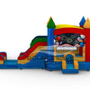 side view of Seasonal Sampler Commercial Econo Inflatable Panel Combo displaying a patriotic Fourth of July themed panel with fireworks, balloons, and American flags. The inflatable includes colorful castle-like towers, dual slides in red and green, and a bounce house area. The structure is set on a blue base with yellow entrance steps and features multiple spires in various colors.
