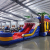 Wide-angle view of Shark Attack Dual Lane Commercial Inflatable Combo set up in a large indoor warehouse. The inflatable features a giant gray shark head with open mouth, dual slides in red and yellow, and inflatable palm trees. A blue splash pool area is visible at the base. The warehouse setting shows metal roofing, bright lighting, and various other inflatable products in the background.