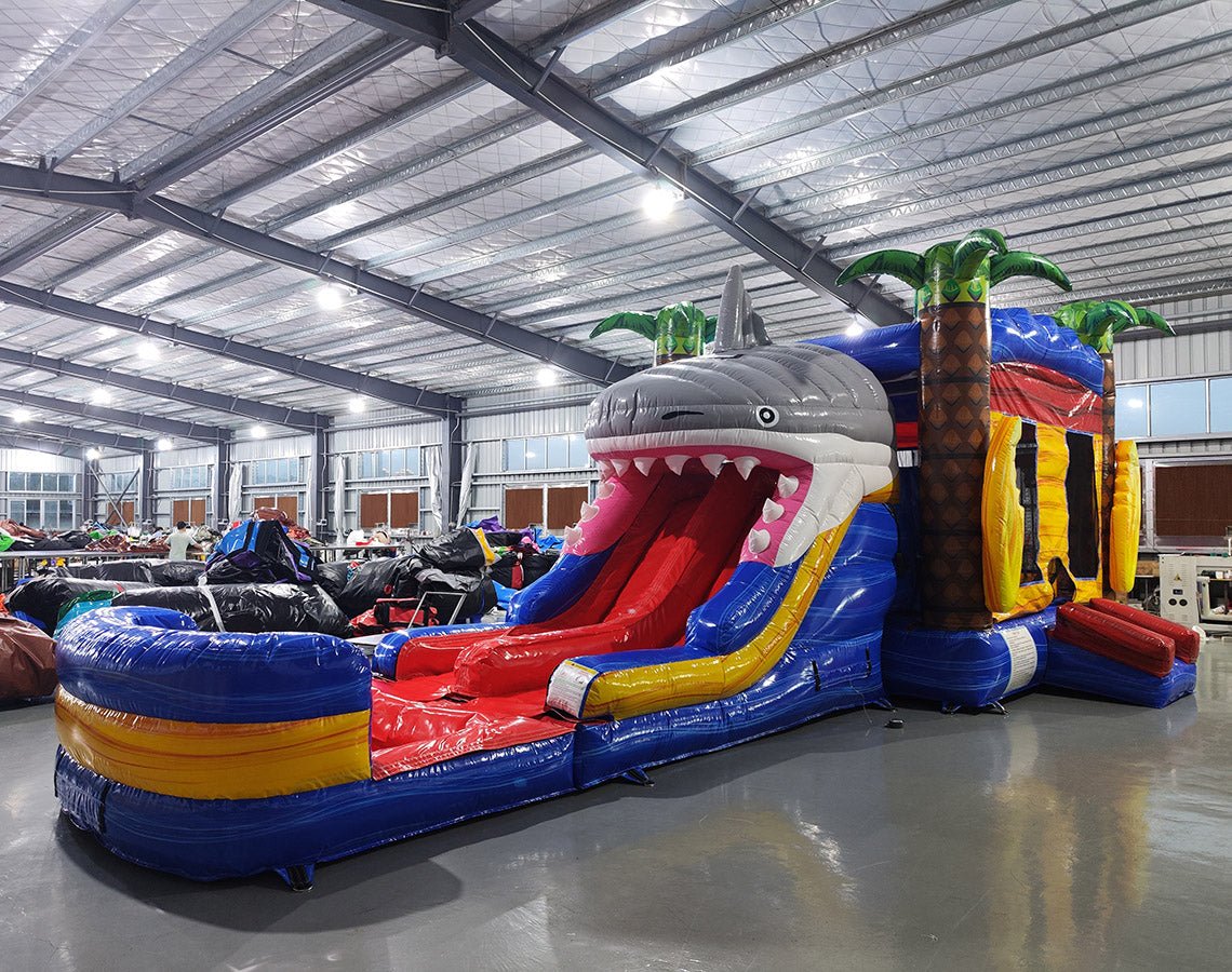 Wide-angle view of Shark Attack Dual Lane Commercial Inflatable Combo set up in a large indoor warehouse. The inflatable features a giant gray shark head with open mouth, dual slides in red and yellow, and inflatable palm trees. A blue splash pool area is visible at the base. The warehouse setting shows metal roofing, bright lighting, and various other inflatable products in the background.