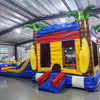 Side view of Shark Attack Dual Lane Commercial Inflatable Combo (Wet/Dry) showing the full length of the inflatable. Features a large gray shark head with open mouth, dual slides in red and yellow emerging from the mouth, and inflatable palm trees. A yellow bounce house area with mesh windows is visible on the side. Set in a spacious warehouse with concrete flooring and various other inflatable products and equipment in the background.