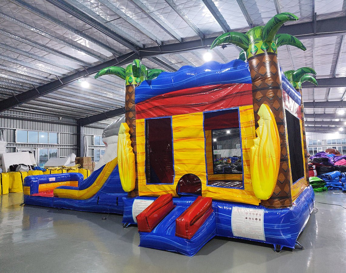 Side view of Shark Attack Dual Lane Commercial Inflatable Combo (Wet/Dry) showing the full length of the inflatable. Features a large gray shark head with open mouth, dual slides in red and yellow emerging from the mouth, and inflatable palm trees. A yellow bounce house area with mesh windows is visible on the side. Set in a spacious warehouse with concrete flooring and various other inflatable products and equipment in the background.