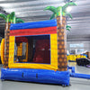 Rear view of Shark Attack Dual Lane Commercial Inflatable Combo (Wet/Dry) featuring a large gray shark head with open mouth and teeth. The structure includes dual slides emerging from the shark's mouth, one red and one yellow, leading to a blue splash pool. Inflatable palm trees flank the sides. Set up in a large indoor warehouse with metal roofing, bright lighting, and various other inflatable products visible in the background.