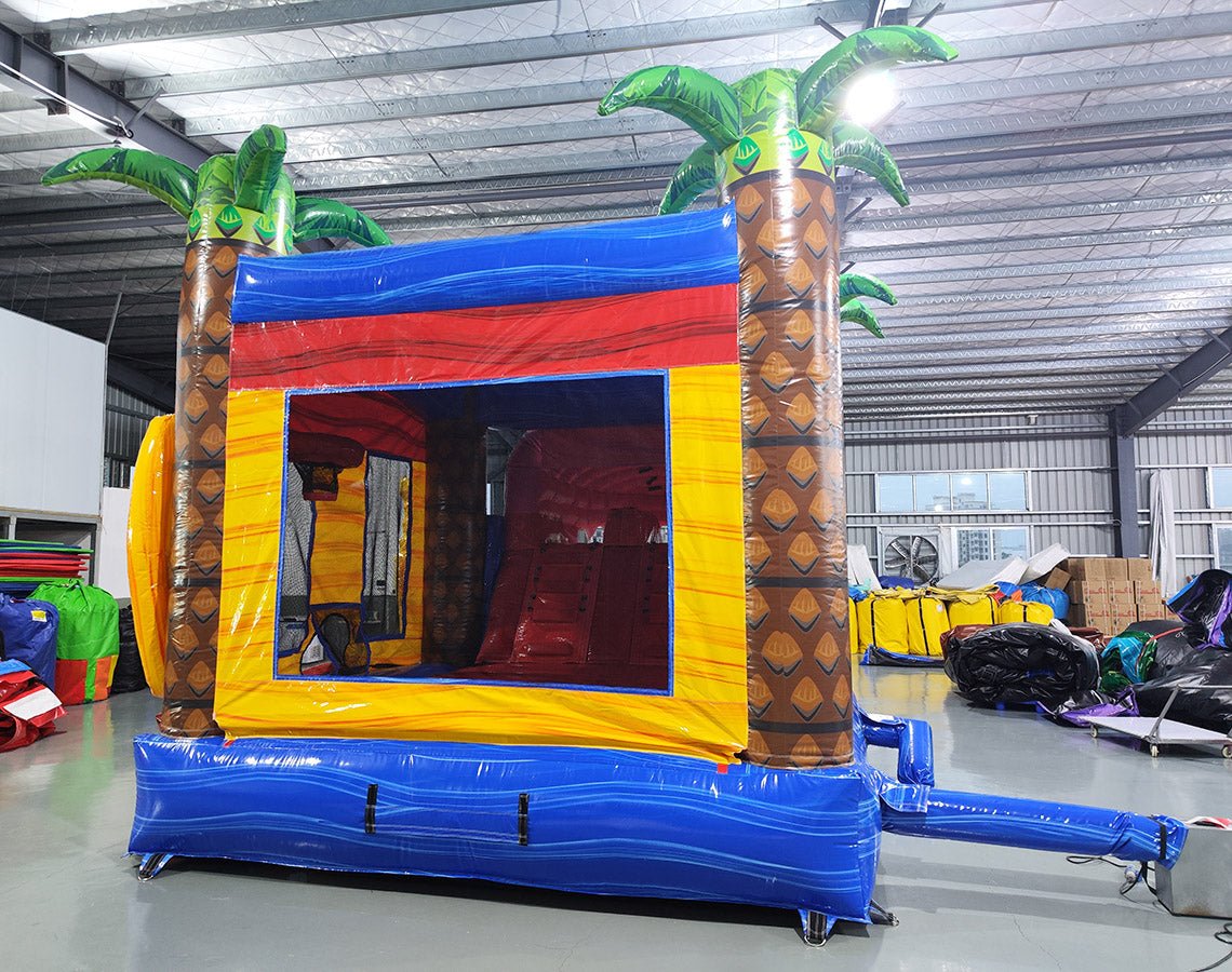 Rear view of Shark Attack Dual Lane Commercial Inflatable Combo (Wet/Dry) featuring a large gray shark head with open mouth and teeth. The structure includes dual slides emerging from the shark's mouth, one red and one yellow, leading to a blue splash pool. Inflatable palm trees flank the sides. Set up in a large indoor warehouse with metal roofing, bright lighting, and various other inflatable products visible in the background.