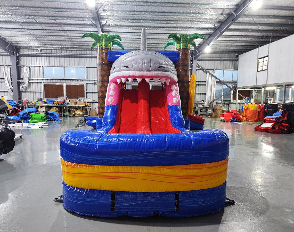 Close-up front view of Shark Attack Dual Lane Commercial Inflatable Combo (Wet/Dry) focusing on the shark head entrance. The large gray shark head has an open mouth with white teeth, red interior, and eyes. Dual slides, one red and one yellow, emerge from the mouth. Inflatable palm trees are visible on either side. The image shows the detailed design of the shark's features against a warehouse backdrop.