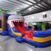 Front view of Shark Attack Dual Lane Commercial Inflatable Combo showing the entrance area. Features a large gray shark head with open mouth and teeth, dual slides in red and yellow emerging from the mouth, and inflatable palm trees. A blue bounce house area with mesh windows is visible on the side. Set in a warehouse with concrete flooring and various other inflatable products and equipment visible in the background.