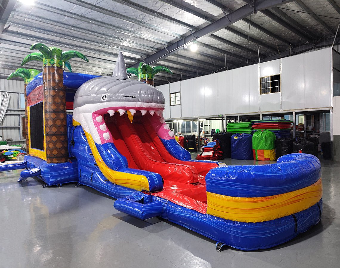 Front view of Shark Attack Dual Lane Commercial Inflatable Combo showing the entrance area. Features a large gray shark head with open mouth and teeth, dual slides in red and yellow emerging from the mouth, and inflatable palm trees. A blue bounce house area with mesh windows is visible on the side. Set in a warehouse with concrete flooring and various other inflatable products and equipment visible in the background.