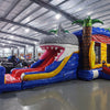 Three-quarter view of Shark Attack Dual Lane Commercial Inflatable Combo in a spacious warehouse. The inflatable displays a large gray shark head with open mouth, dual slides in red and yellow, and inflatable palm trees. A blue bounce house area with mesh windows is visible on the side. Various other inflatable products and warehouse equipment can be seen in the background.