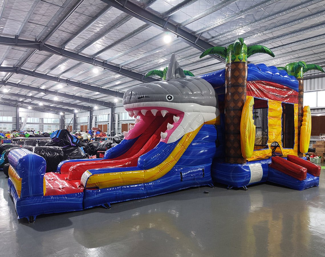 Three-quarter view of Shark Attack Dual Lane Commercial Inflatable Combo in a spacious warehouse. The inflatable displays a large gray shark head with open mouth, dual slides in red and yellow, and inflatable palm trees. A blue bounce house area with mesh windows is visible on the side. Various other inflatable products and warehouse equipment can be seen in the background.