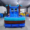 Front view of South Beach Dual Lane Commercial Inflatable Combo (Wet/Dry) featuring a tropical theme with inflatable palm trees. The structure has dual blue slides, a turquoise bounce house area with mesh windows, and a blue splash pool base. Large inflatable sunglasses with beach scenes adorn the top. Set up in a large indoor warehouse with metal roofing and various equipment visible in the background.