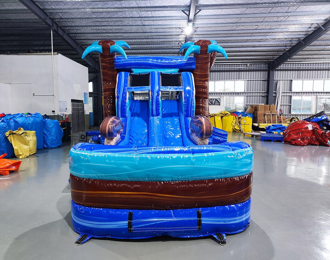 Front view of South Beach Dual Lane Commercial Inflatable Combo (Wet/Dry) featuring a tropical theme with inflatable palm trees. The structure has dual blue slides, a turquoise bounce house area with mesh windows, and a blue splash pool base. Large inflatable sunglasses with beach scenes adorn the top. Set up in a large indoor warehouse with metal roofing and various equipment visible in the background.