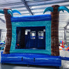 Rear view of South Beach Dual Lane Commercial Inflatable Combo (Wet/Dry) featuring a tropical theme with inflatable palm trees and large sunglasses. The structure includes dual blue slides, a turquoise bounce house area with mesh windows, and a blue splash pool base. Set up in a large indoor warehouse with metal roofing and various equipment visible in the background.