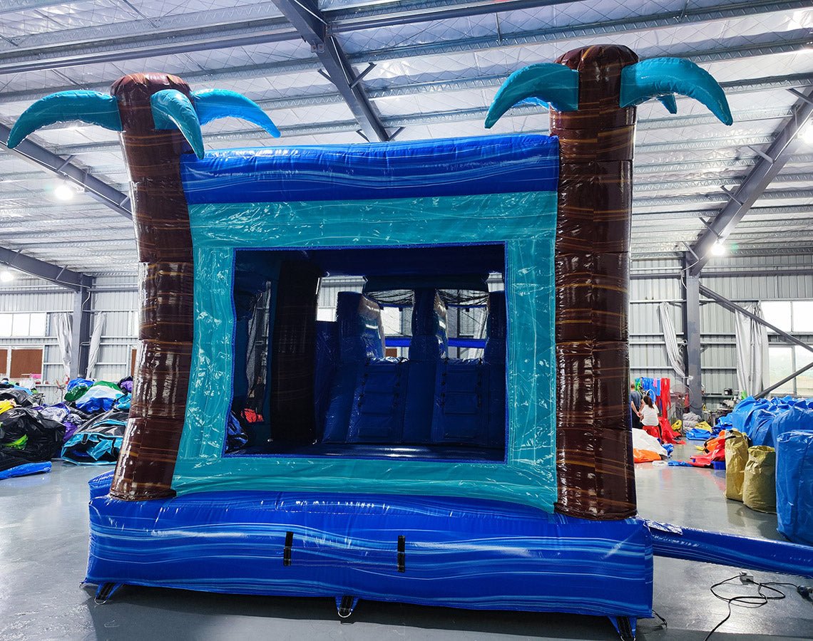 Rear view of South Beach Dual Lane Commercial Inflatable Combo (Wet/Dry) featuring a tropical theme with inflatable palm trees and large sunglasses. The structure includes dual blue slides, a turquoise bounce house area with mesh windows, and a blue splash pool base. Set up in a large indoor warehouse with metal roofing and various equipment visible in the background.