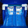 Inter view of South Beach Dual Lane Commercial Inflatable Combo (Wet/Dry) showing dual blue slides, brown palm tree pillars, and a turquoise bounce house. Large inflatable sunglasses with beach scenes top the structure. The warehouse setting includes metal roofing and various other inflatable products, showcasing its commercial application.