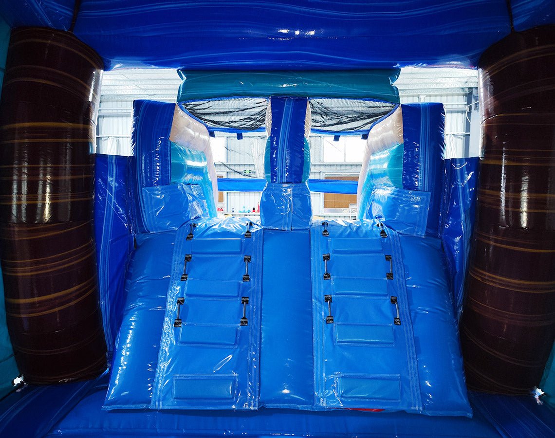 Inter view of South Beach Dual Lane Commercial Inflatable Combo (Wet/Dry) showing dual blue slides, brown palm tree pillars, and a turquoise bounce house. Large inflatable sunglasses with beach scenes top the structure. The warehouse setting includes metal roofing and various other inflatable products, showcasing its commercial application.