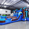 Three-quarter view of South Beach Dual Lane Commercial Inflatable Combo (Wet/Dry) set up in a warehouse. The inflatable features dual blue slides, brown palm tree pillars, a turquoise bounce house, and a blue splash pool base. Large sunglasses with beach scenes top the structure. Various other inflatable products and equipment are visible in the background, showcasing its commercial setting.